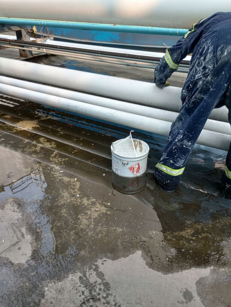 A painter standing in a shallow pool of dirty water applies a protective coating to a coated steel pipe at a liquid terminal.